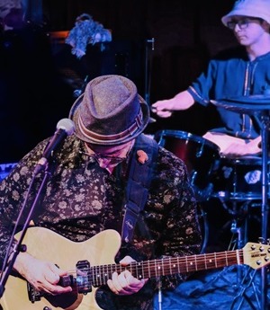 Two members of the band Sloth Broth perform on stage. One man in the foreground plays a guitar and one man in the background plays drums.
