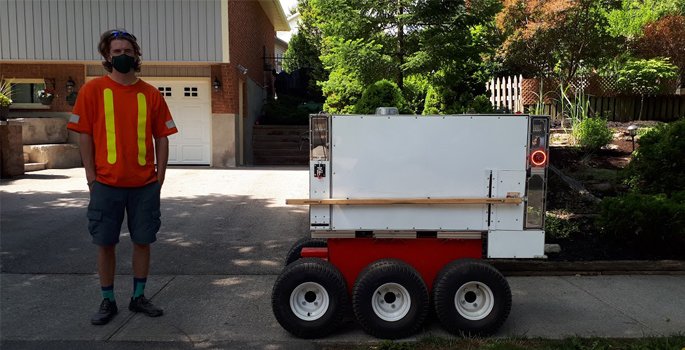 Staff person on sidewalk standing beside simple robot with wheels